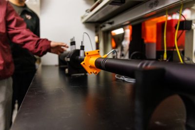 A long plastic tube with fittings is set up on a lab bench, with researchers standing nearby.